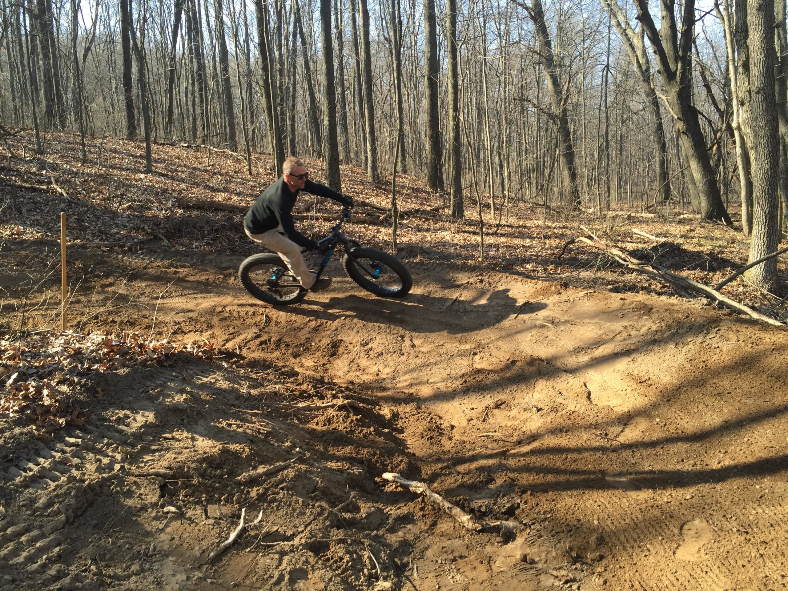 A person riding a mountain bike on a dirt trail in a forest. The rider, wearing a black sweater and sunglasses, leans into a curve on a sandy path surrounded by trees and fallen leaves. The scene captures the excitement of mountain biking in a natural setting. DTE Energy Foundation Trail mountain bike trail.