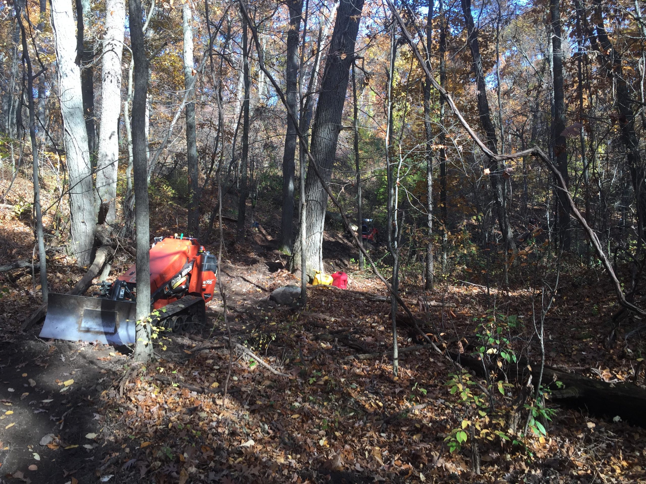 A red tractor with a front loader is parked on a dirt path in a wooded area with autumn foliage. The ground is covered in fallen leaves, and several trees surround the scene, creating a natural environment. In the background, hints of equipment and tools can be seen, suggesting active work in the forest. DTE Energy Foundation Trail mountain bike trail.