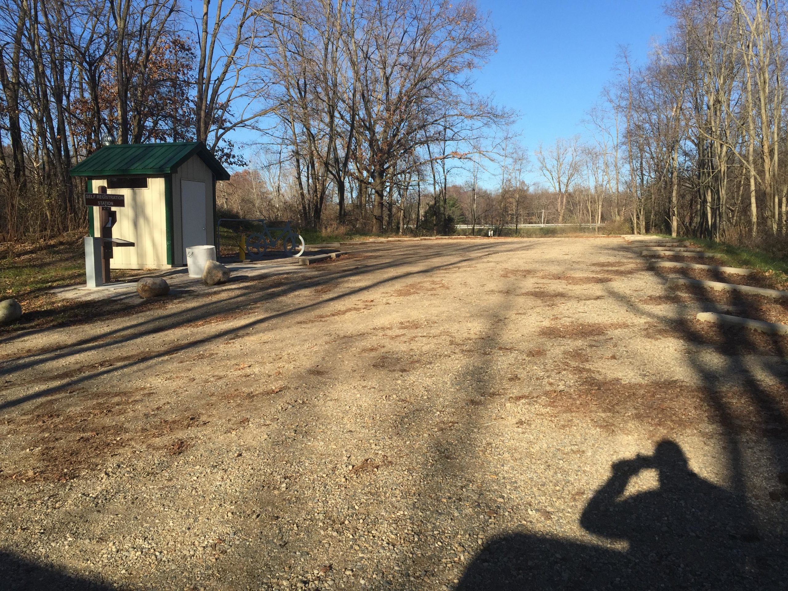 A gravel path in a park setting with a small building featuring a green roof and signage. Surrounding trees are bare, indicating late autumn or early winter. The area is calm and mostly empty, with a few large rocks positioned nearby. A shadow of a person holding a camera is visible in the foreground. DTE Energy Foundation Trail mountain bike trail.