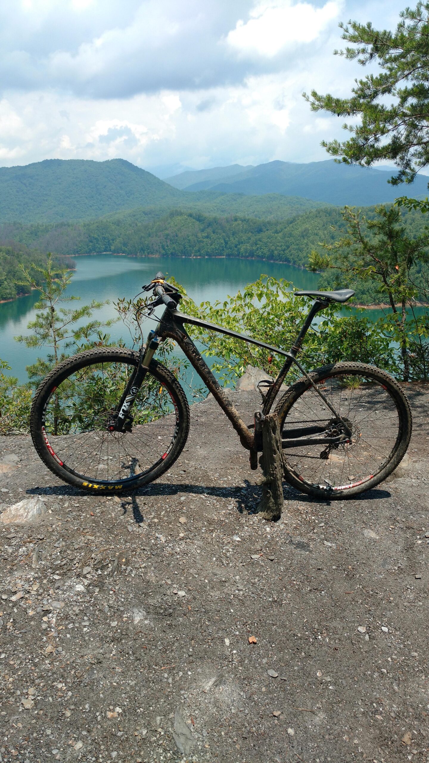 Specialized S-WORKS STUMPJUMPER HT: A mountain bike resting on a rocky surface overlooking a calm lake surrounded by green hills and mountains under a partly cloudy sky. The bike is muddy, indicating recent use on a rugged trail.