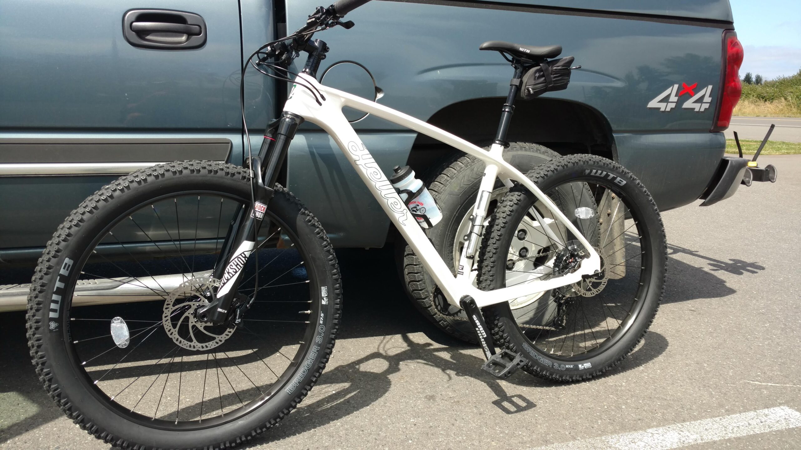 Heller Shagamaw: A white mountain bike with oversized tires is parked next to a blue pickup truck. The bike features a front suspension fork and a water bottle holder mounted on its frame. The truck has a "4x4" emblem on its rear. Sunlight casts a shadow of the bike on the asphalt surface.