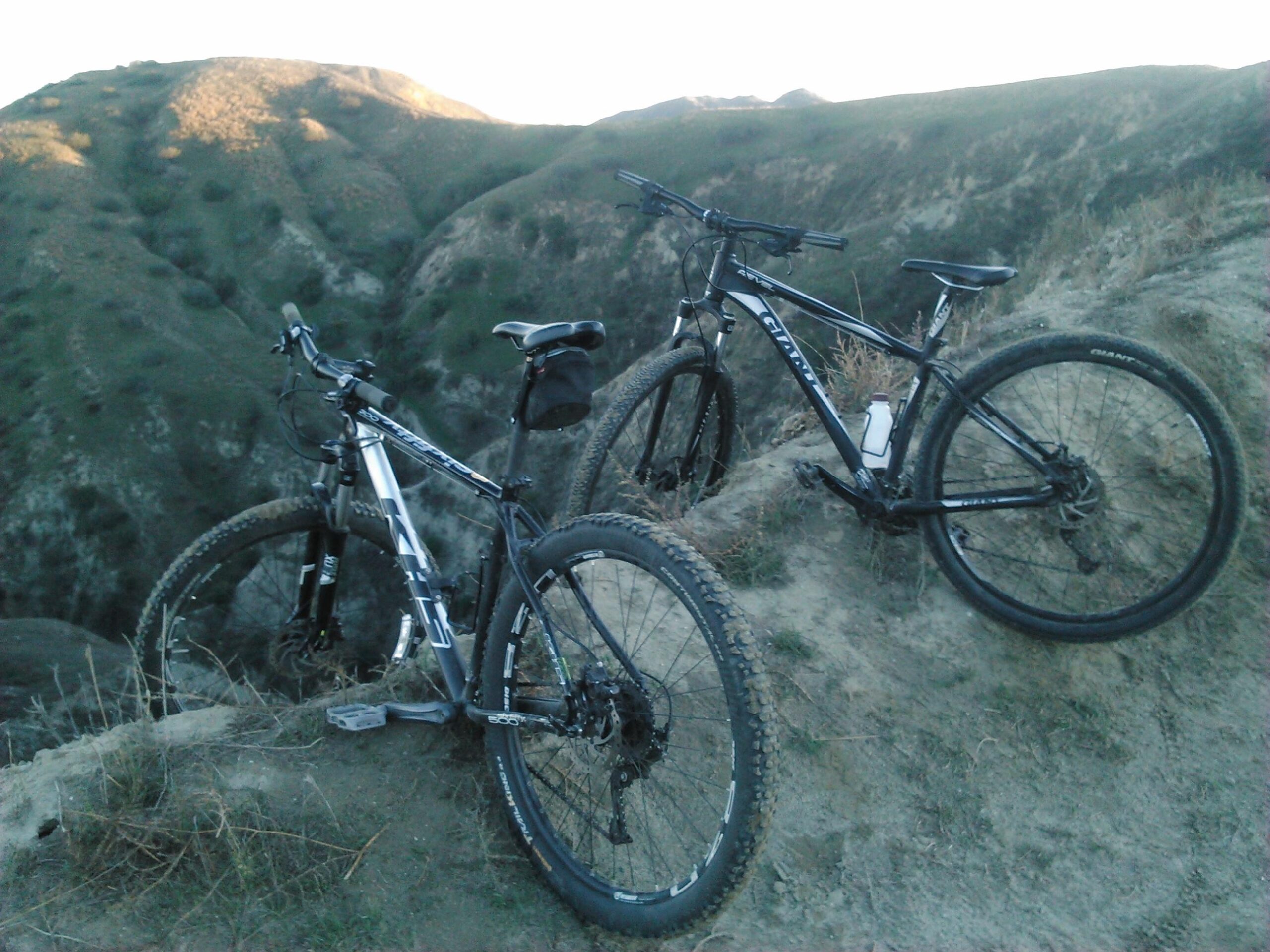KHS SixFifty 600: Two mountain bikes resting on a dirt hill with a background of rolling green hills under a clear sky. One bike is silver with black accents and a small storage bag, while the other is predominantly black with white details.