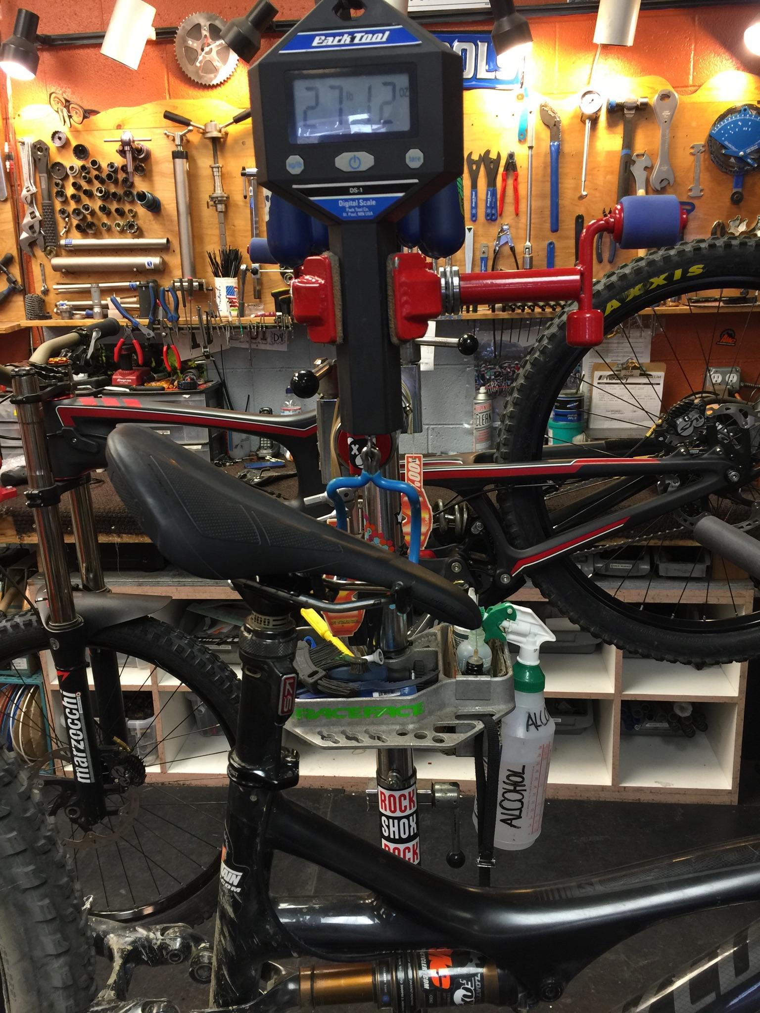 Specialized Stumpjumper: A digital scale displaying 27.13 ounces measures a mountain bike suspended from a workstation. The background features various tools and bike parts organized on a wall, with a bottle of cleaning solution labeled "ALCOHOL" on the bike stand.