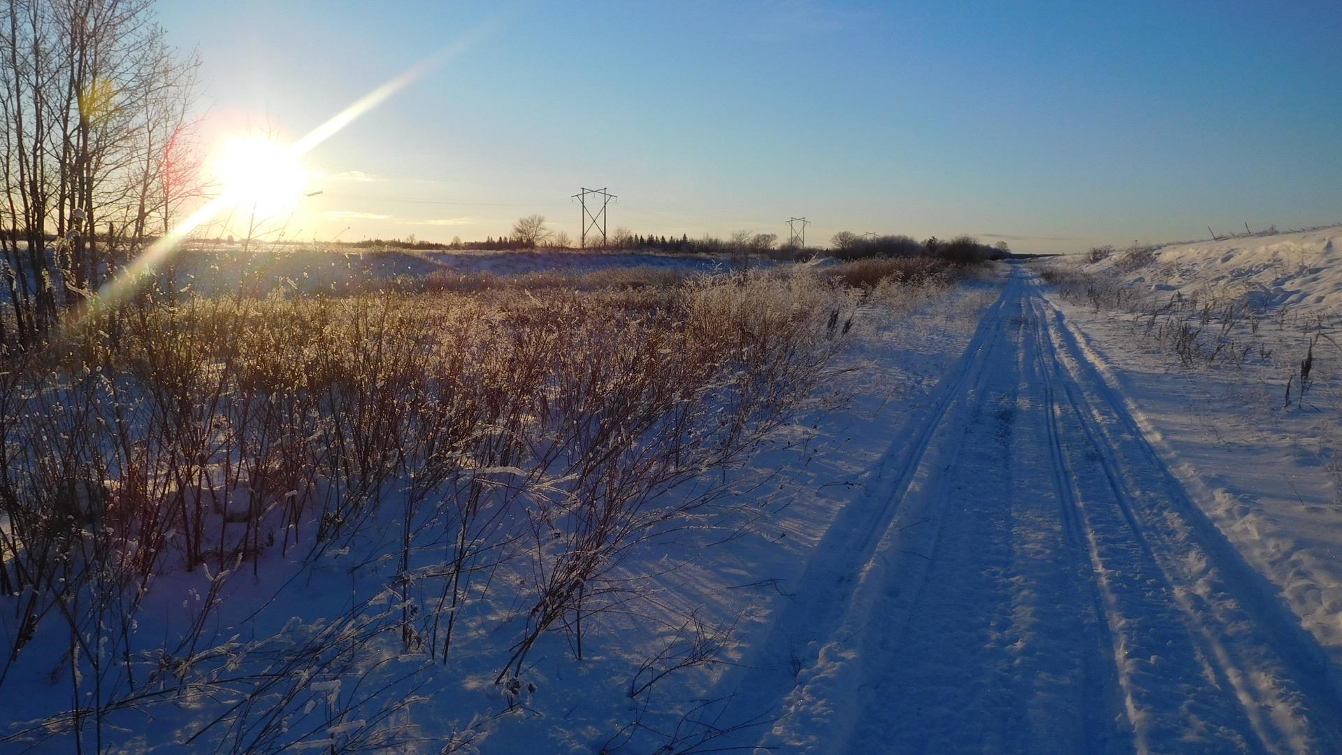 A snow-covered path winding through a serene landscape, with the sun setting in the background and casting a warm glow. Bare trees and frosty grasses line the sides of the path, and power lines are visible in the distance against a clear blue sky. Seven Sisters Dam to Whiteshell Park gate @ HWY #307 mountain bike trail.
