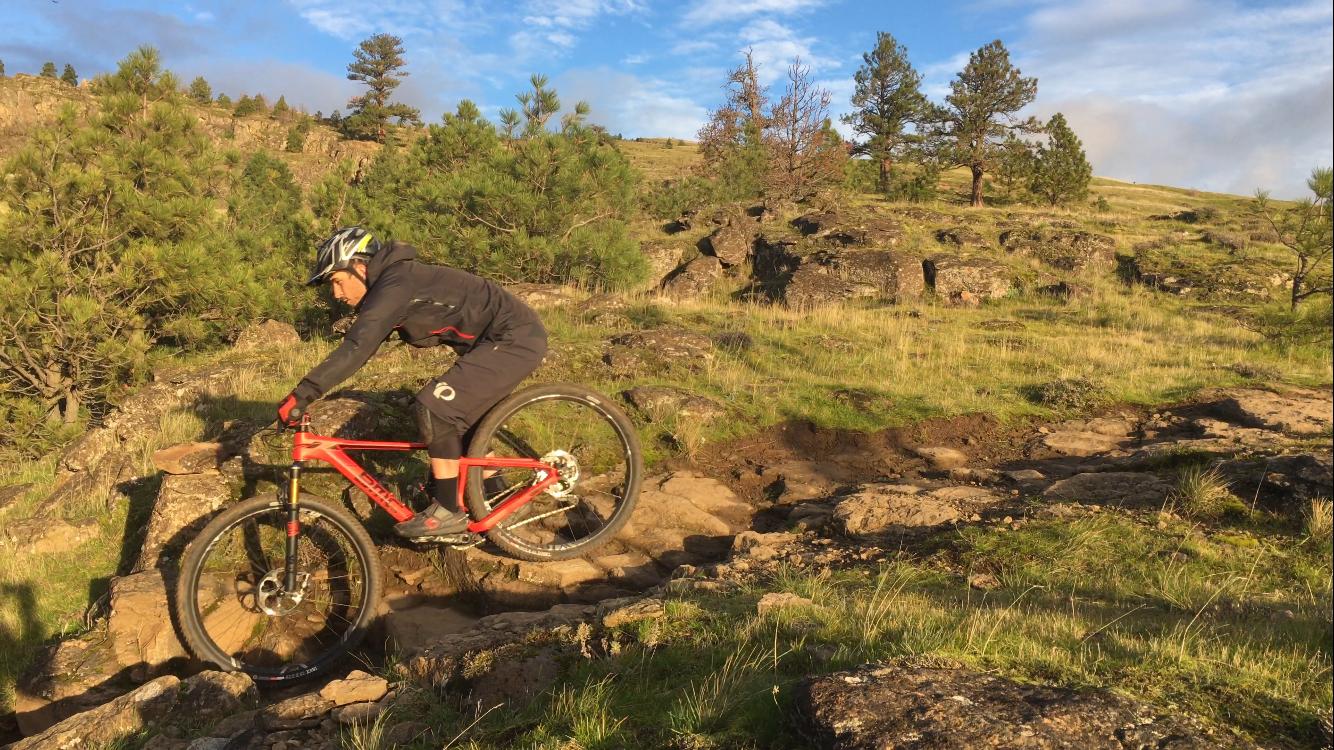 A mountain biker navigating a rocky trail surrounded by greenery and trees, with a partly cloudy blue sky in the background. The biker is in action, balancing on the bike while lifting the front wheel off the ground. Syncline mountain bike trail.