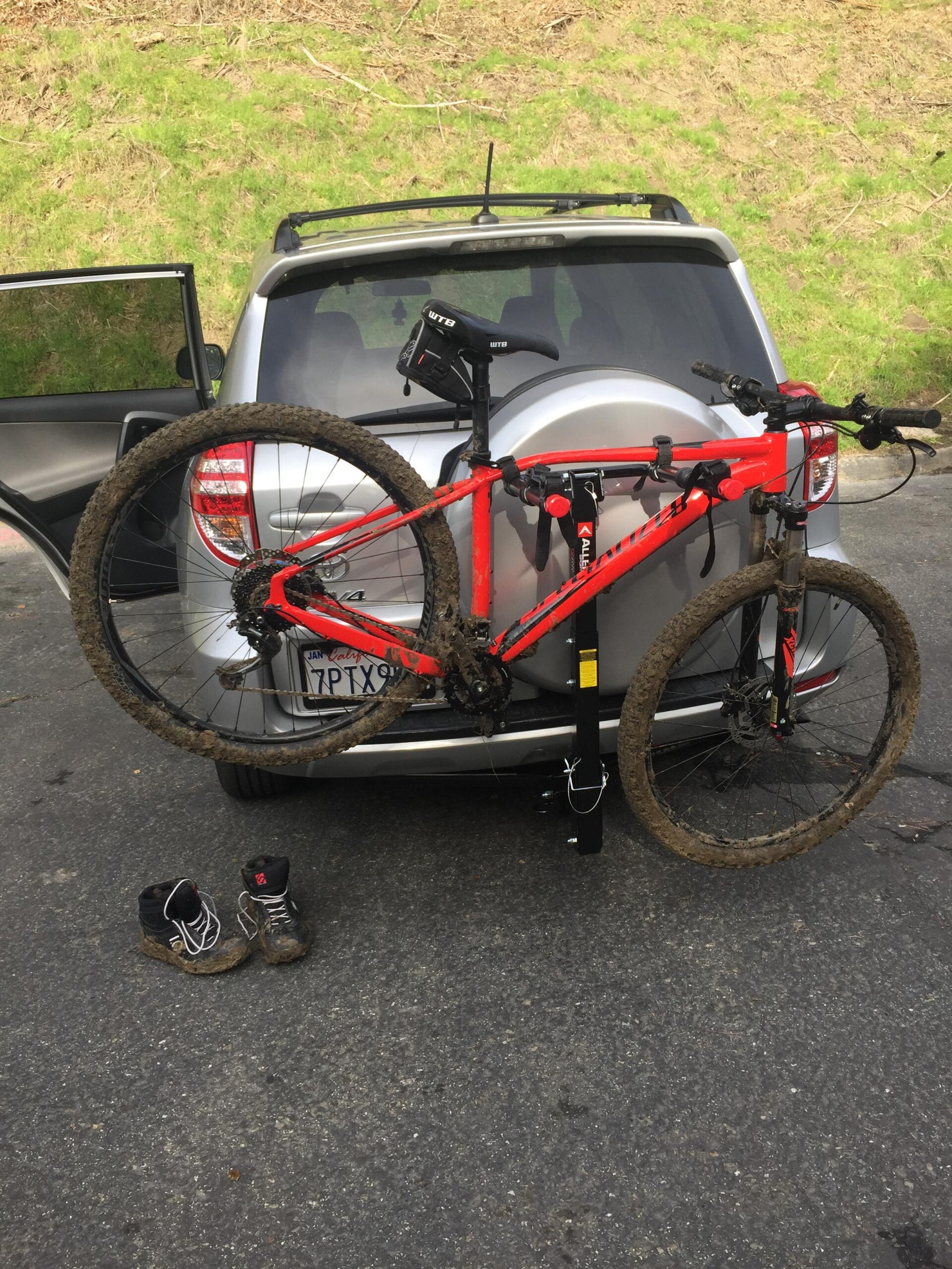 Specialized Rockhopper Comp 29: A red mountain bike mounted on the back of a silver SUV, partially obscured by an open rear door. The bike shows signs of mud, indicating recent use on a trail. A pair of muddy shoes is placed on the ground nearby. In the background, a grassy area is visible.