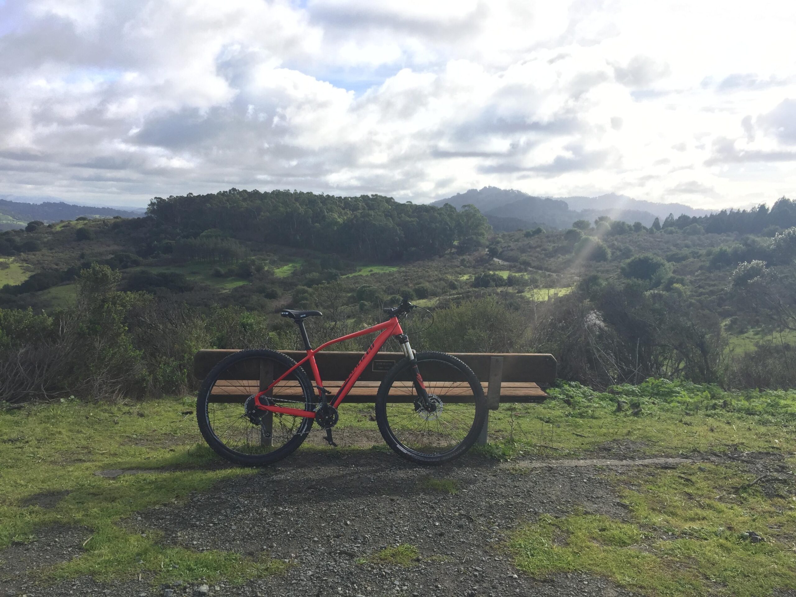 Specialized Rockhopper Comp 29: A bright red mountain bike is parked on a wooden bench overlooking a scenic landscape with rolling hills and patches of greenery under a partly cloudy sky. In the background, distant mountains can be seen.
