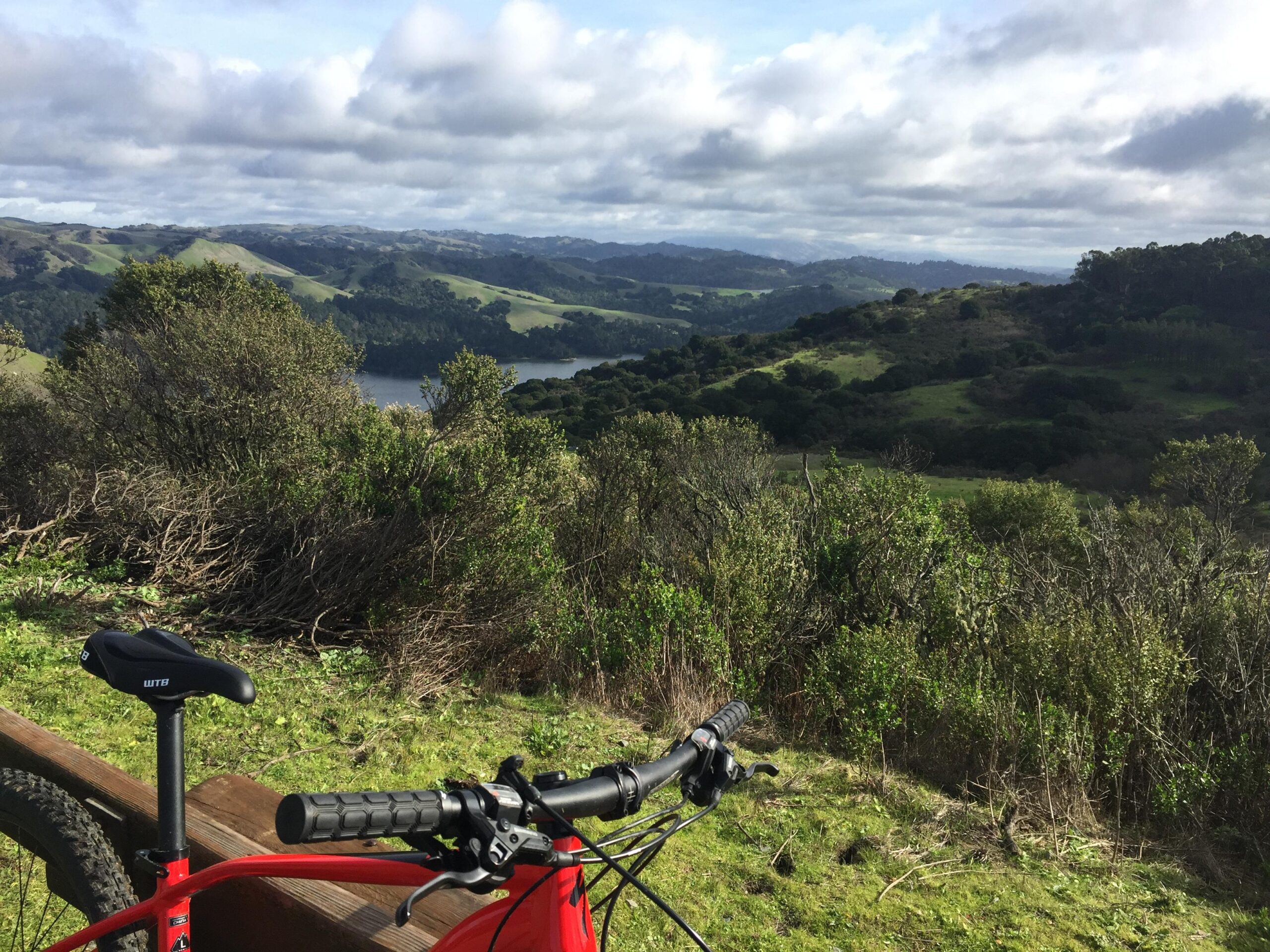 Specialized Rockhopper Comp 29: A mountain bike is propped against a wooden railing, overlooking a scenic landscape with rolling green hills and a lake in the distance. The sky is partly cloudy, adding a soft light to the serene outdoor setting.