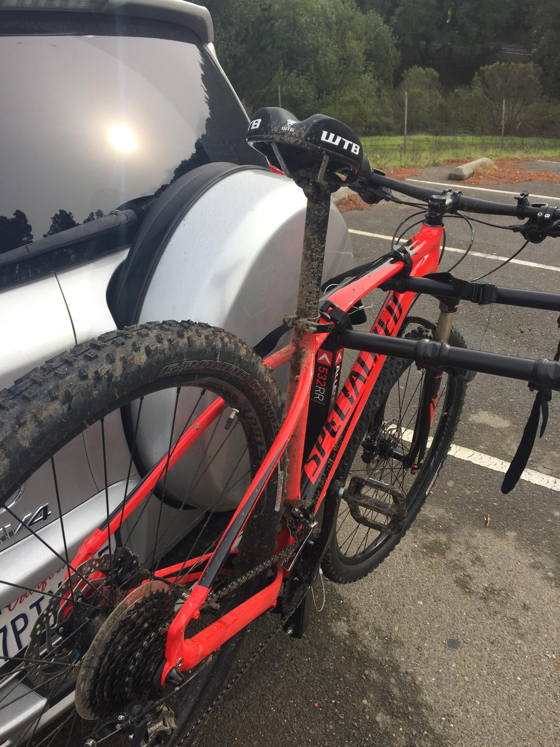 Specialized Rockhopper Comp 29: A red mountain bike with a muddy frame is mounted on the back of a silver SUV in a parking area surrounded by trees. The bike's saddle features the WTB logo, and the rear wheel is visible alongside the vehicle's rear.