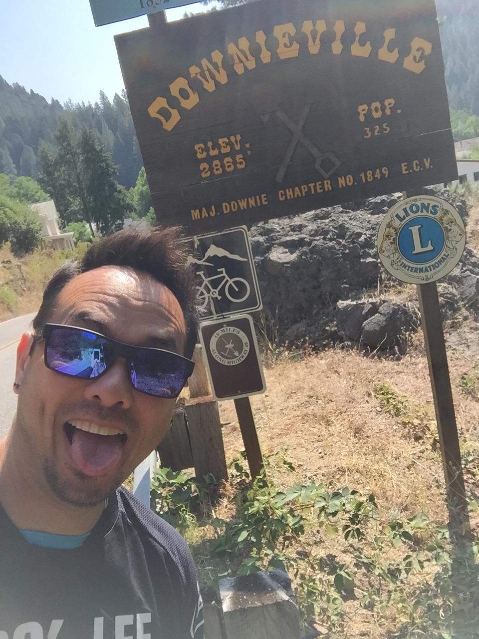 Specialized Stumpjumper: A person wearing sunglasses, smiling and sticking out their tongue, stands in front of a weathered wooden sign that reads "Downieville" with elevation and population information. The background features trees and rocky terrain, indicating a rural setting. The sign also has symbols related to biking and a Lions Club emblem.