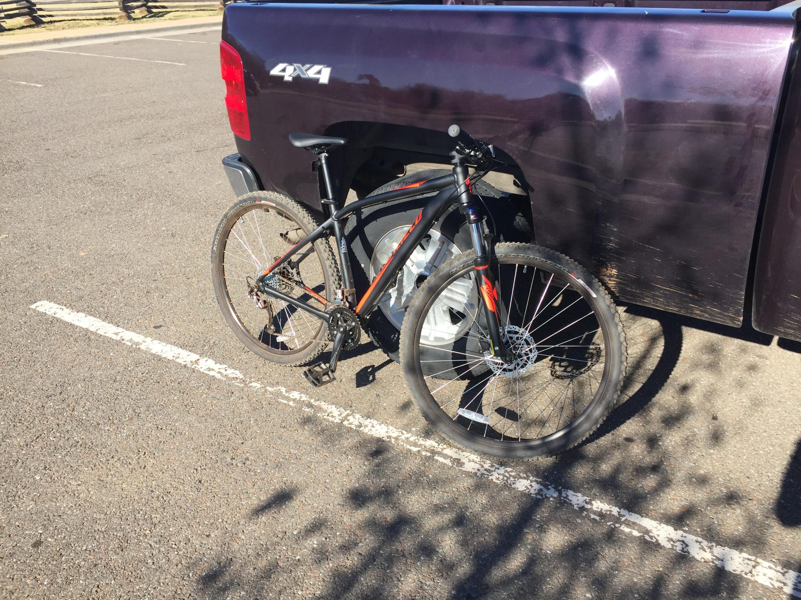 Specialized Rockhopper Sport 29: A black mountain bike with orange accents parked next to a purple pickup truck in a parking lot. The bike is secured to the truck, with the sun casting shadows on the ground.