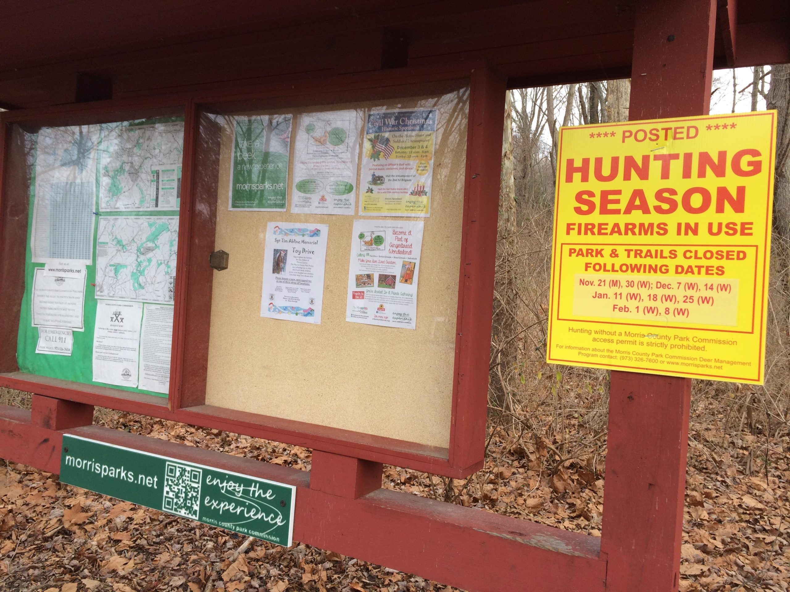 Information board at a park featuring community announcements, maps, and a yellow sign indicating hunting season dates and safety reminders. The sign informs visitors about park and trail closures on specific dates due to firearms being in use. The board is surrounded by bare trees and leaf litter. Lewis Morris mountain bike trail.