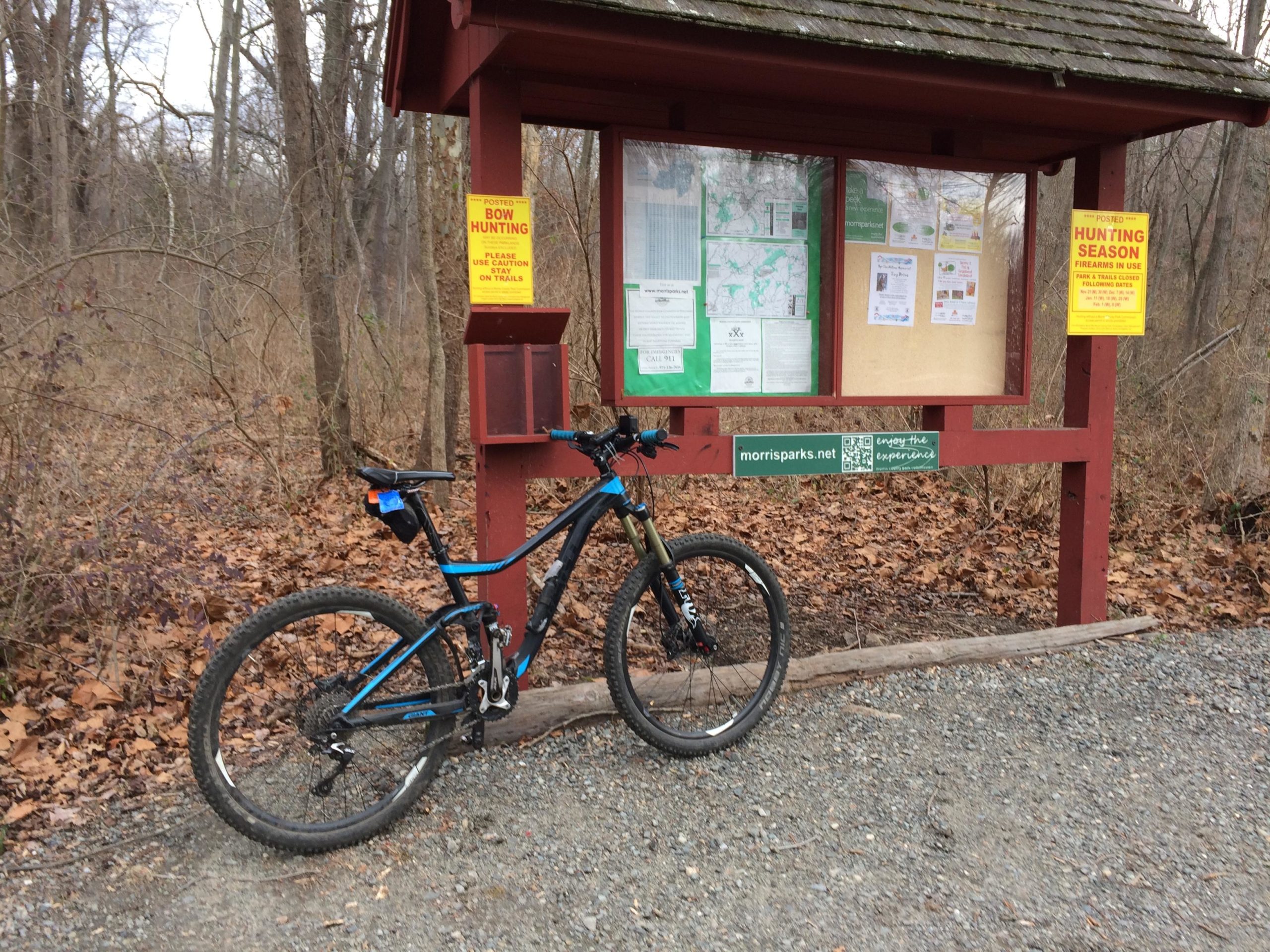 A mountain bike is leaning against an information kiosk at a trailhead in a wooded area. The kiosk features various signs, including warnings about bow hunting and hunting season, and is surrounded by fallen leaves and trees in a natural setting. Lewis Morris mountain bike trail.