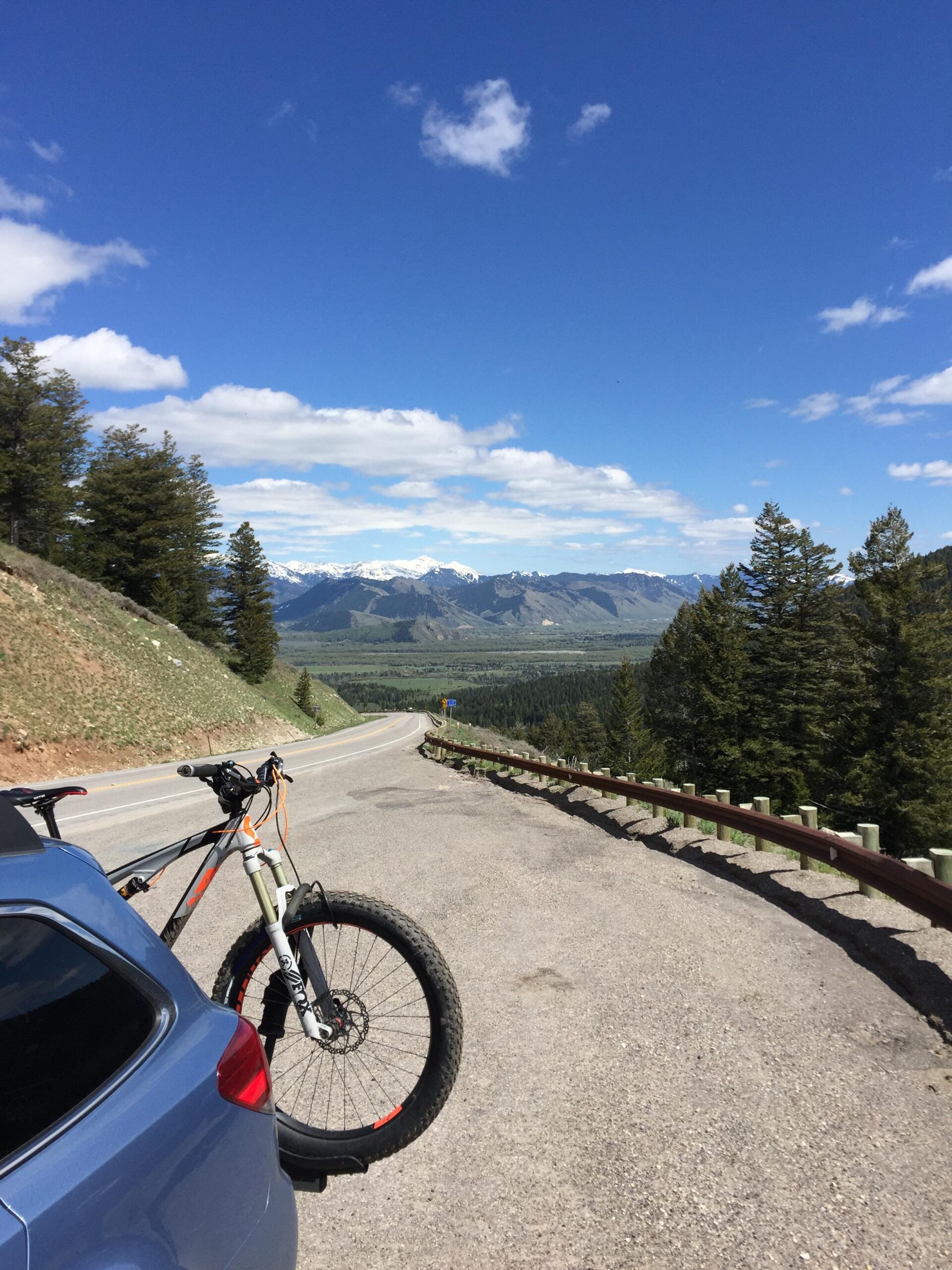 Scott Genius Plus: A scenic view from a roadside overlook, featuring a mountain bike mounted on the back of a blue car. In the background, snow-capped mountains rise against a bright blue sky dotted with clouds. Lush greenery and trees line the road, which curves along a valley below.