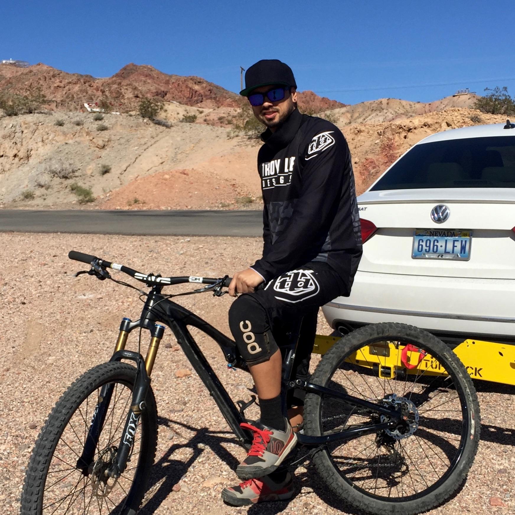 Specialized Stumpjumper: A person wearing a black long-sleeve shirt and shorts is sitting on a mountain bike in a rocky outdoor setting. They are wearing sunglasses, knee pads, and sports shoes. A white car is parked in the background, with mountainous terrain and a clear blue sky overhead.