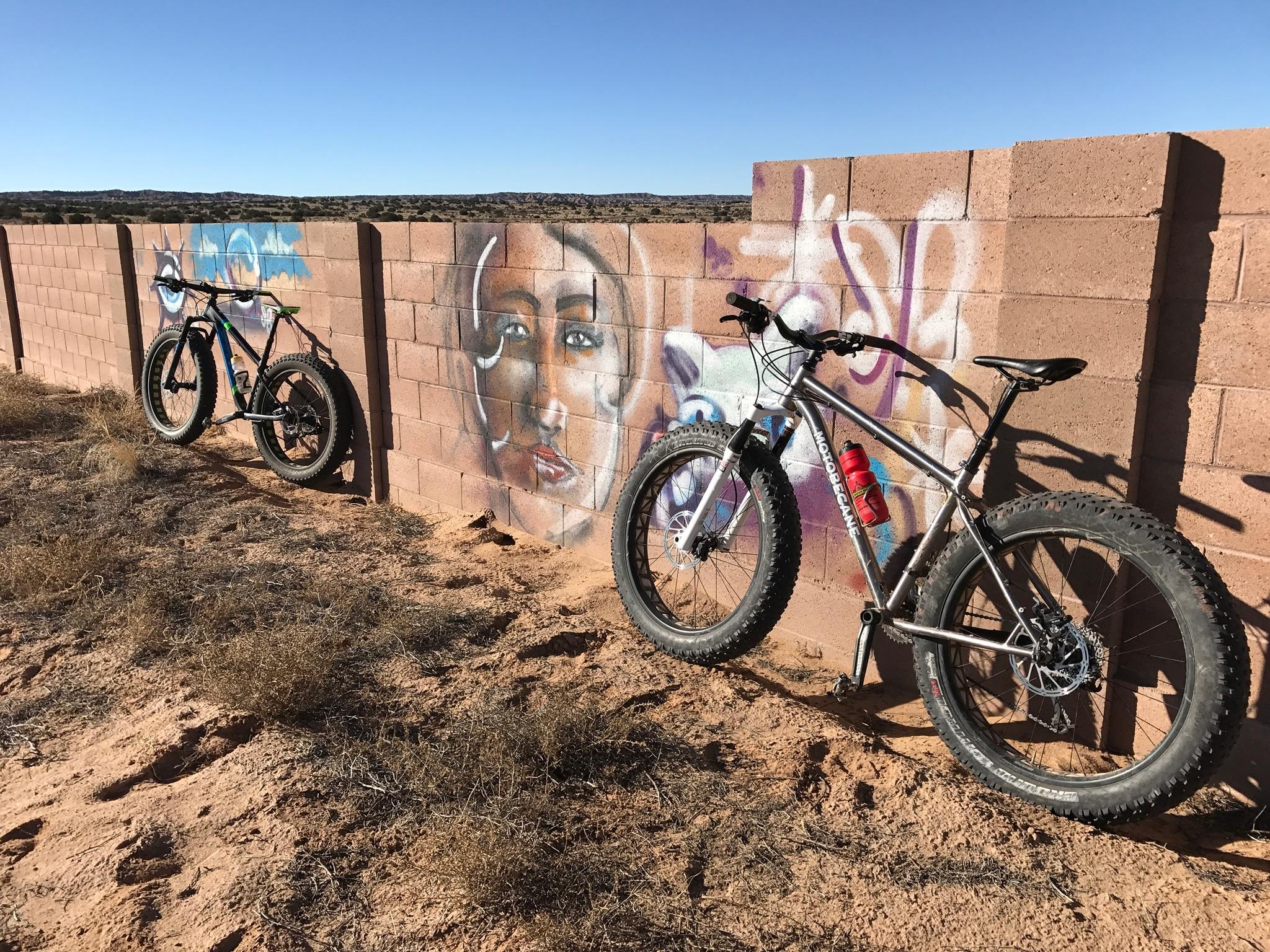 Two mountain bikes are leaning against a textured, artfully painted block wall. The wall features a colorful mural of a face along with abstract designs, set against a clear blue sky. The ground is sandy and sparse, with dried vegetation surrounding the bikes, which have wide tires suited for rough terrain. Super Fat Bike Loop mountain bike trail.