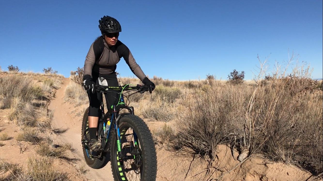 A person riding a mountain bike on a dirt trail surrounded by desert vegetation, wearing a helmet and sporting gear. The landscape features sparse bushes and a clear blue sky, suggesting an outdoor adventure in a natural setting. Super Fat Bike Loop mountain bike trail.