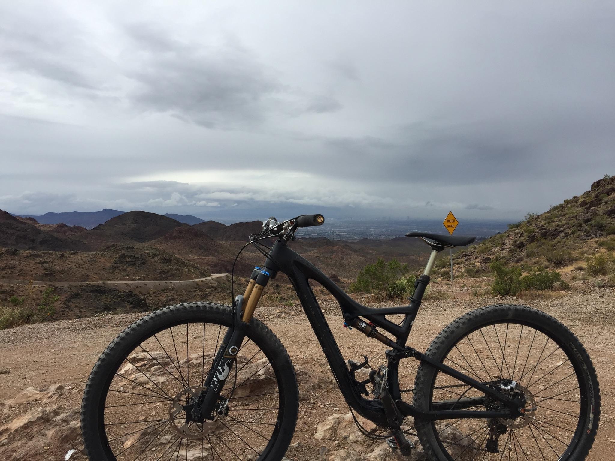 Specialized Stumpjumper: A black mountain bike is positioned on rocky terrain, with a panoramic view of rugged hills and a cloudy sky in the background. A caution sign indicating "Rough Road" is visible in the distance, adding to the outdoor adventure atmosphere.