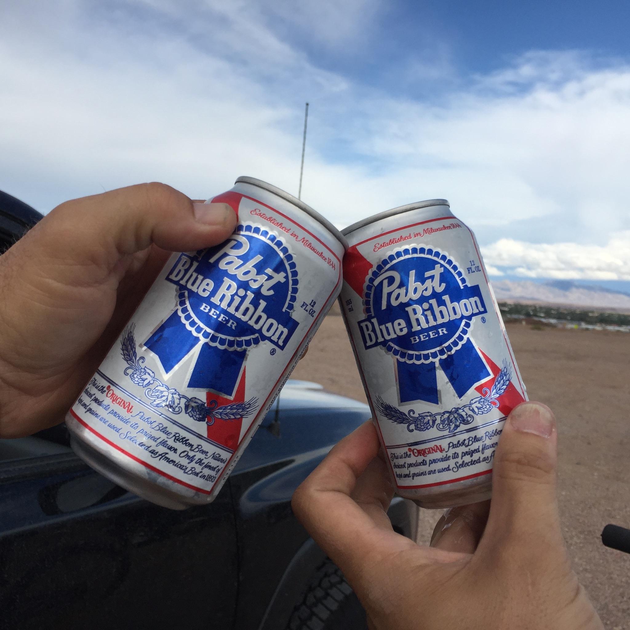 Specialized Stumpjumper: Two hands holding cans of Pabst Blue Ribbon beer, clinking together in a toast against a backdrop of blue skies and landscape.