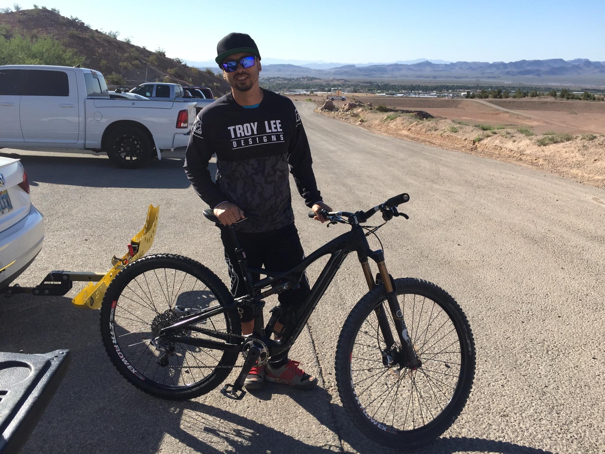 Specialized Stumpjumper: A person stands next to a mountain bike on a dirt road, wearing sunglasses, a black long-sleeve shirt with the logo "Troy Lee Designs," and shorts. In the background, vehicles are parked and mountains are visible in the distance under a clear blue sky.