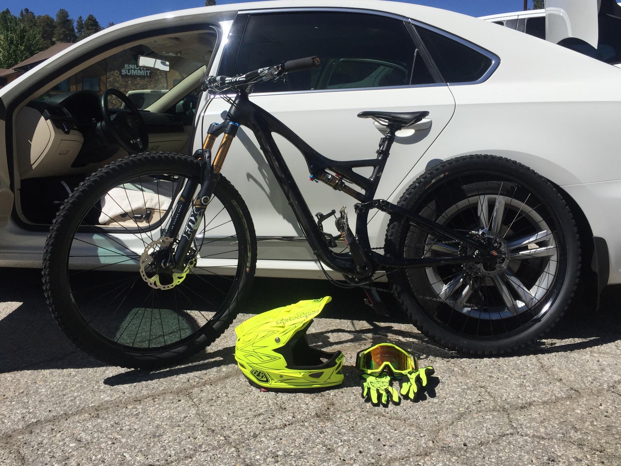 Specialized Stumpjumper: Mountain bike leaning against a white car with the driver's door open. Nearby, a bright yellow helmet, a pair of yellow gloves, and a pair of goggles are placed on the ground. The scene is set against a clear blue sky, hinting at a day of outdoor adventure.