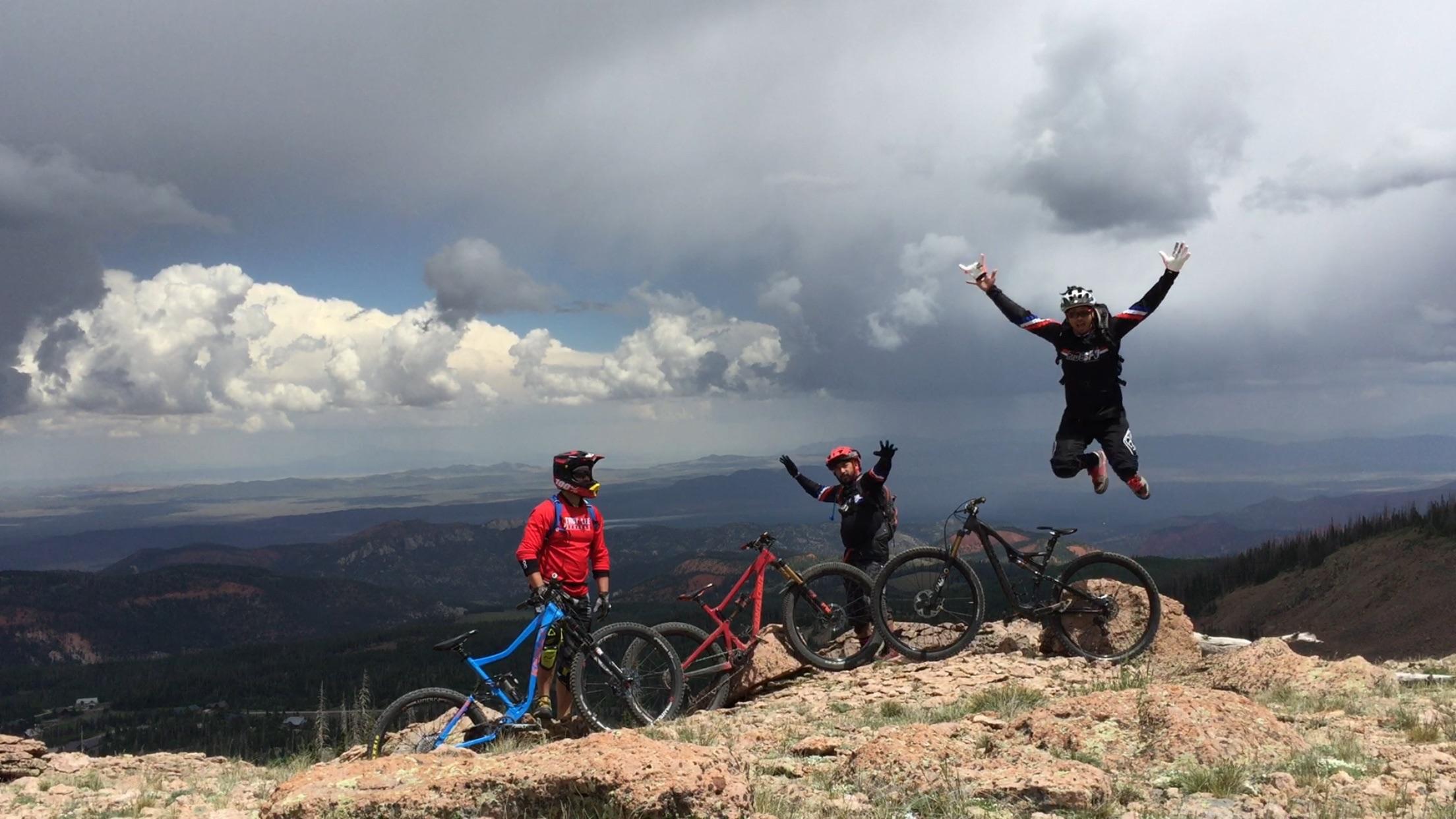 Specialized Stumpjumper: Three mountain bikers pose on a rocky outcrop in a mountainous landscape, with one biker mid-jump, expressing excitement. They are surrounded by their bikes, and the backdrop features dramatic clouds and a distant view of hills and valleys.
