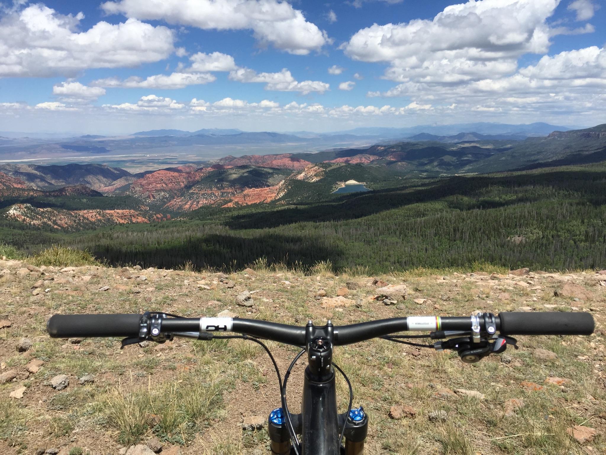 Specialized Stumpjumper: A mountain bike handlebar in the foreground, overlooking a panoramic view of colorful hills and valleys under a partly cloudy sky. The landscape features lush greenery, rocky terrain, and distant mountains, giving a sense of an elevated outdoor adventure.
