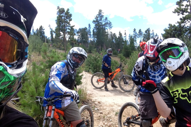 A group of five mountain bikers posing for a selfie on a sunny day in a forested area. They are wearing helmets and protective gear, with their bikes parked nearby. The background features tall pine trees and a dirt trail, indicating an outdoor biking location.