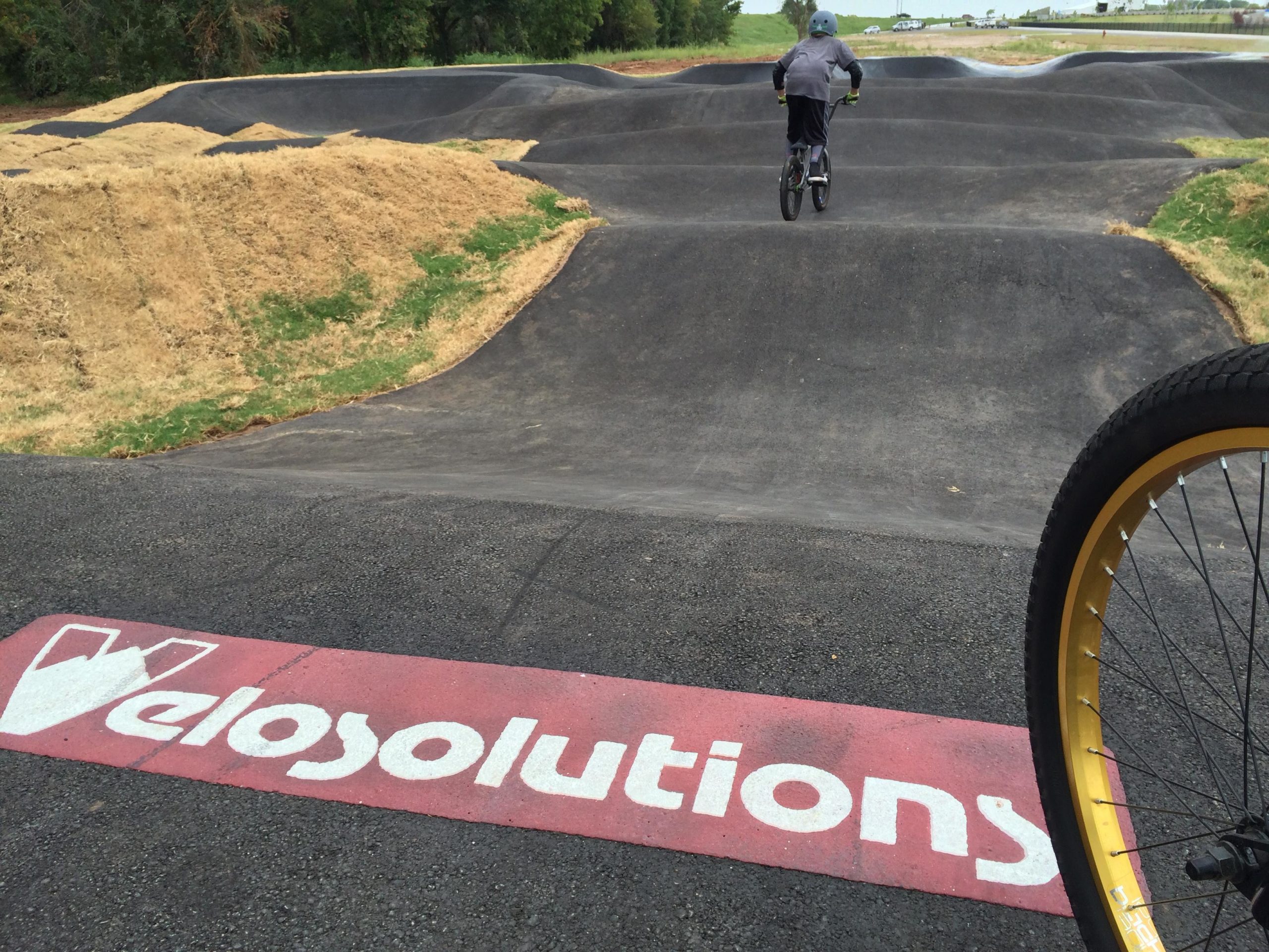 A BMX rider on a pump track, with mounds of dirt and grass surrounding the track. The foreground features a bicycle wheel and a logo that reads "Velosolutions." Riversport Flow Trail mountain bike trail.