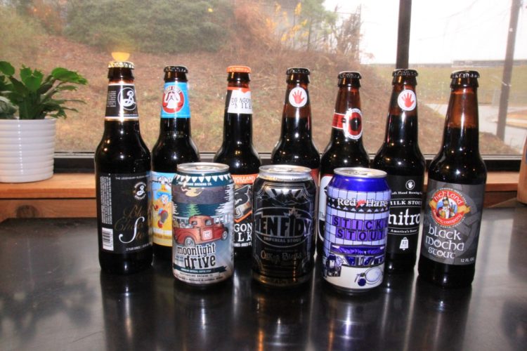 A collection of various beer bottles and cans arranged on a table, with a window in the background showcasing an outdoor view. The assortment includes both dark bottles and colorful cans from different breweries, featuring labels such as "Moonlight Drive," "Ten Fidy," and "Black Mocha Stout." A potted plant is visible in the corner, adding a touch of green to the scene.