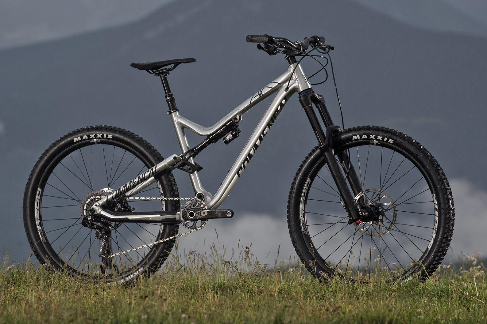 Commencal Meta AM V4: A silver mountain bike positioned on grass, with a scenic mountainous background. The bike features wide tires with Maxxis branding, a prominent front suspension fork, and a sleek frame design. The setting suggests an outdoor adventure environment.