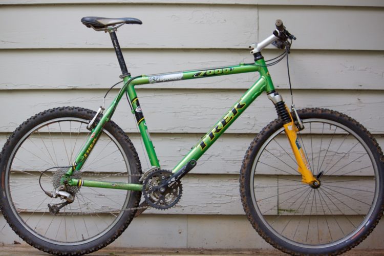An image of a green Trek mountain bike leaning against a light-colored wooden wall. The bike features thick, knobby tires and a suspension fork with an orange accent. The drivetrain includes a visible chain and a rear derailleur. The bike's seat is raised, and the handlebars are equipped with grips and brake levers.