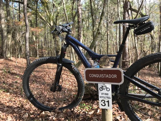 A mountain bike parked next to a trail sign that reads "Conquistador" indicating an off-road bicycle trail numbered 31, surrounded by tall trees and fallen leaves in a wooded area. San Felasco Hammock Preserve mountain bike trail.