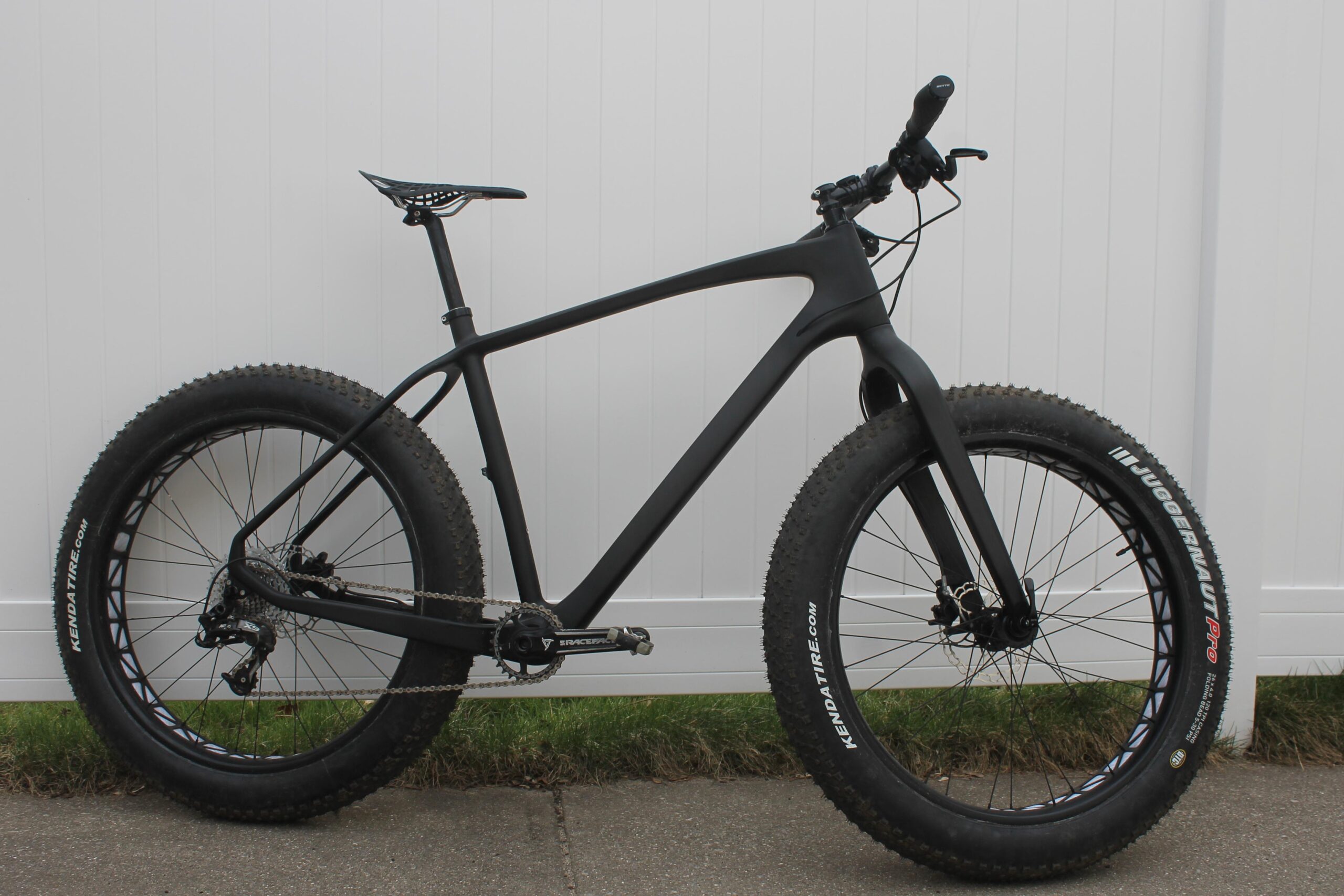 Lamere Carbon Fat Bike: A black fat bike with wide tires is positioned against a white wooden fence. The bike features a sleek frame, a sporty saddle, and a chain system. The tires are knobby and designed for off-road use, highlighting the bike's versatility for various terrains. The setting includes a patch of grass and a paved surface.
