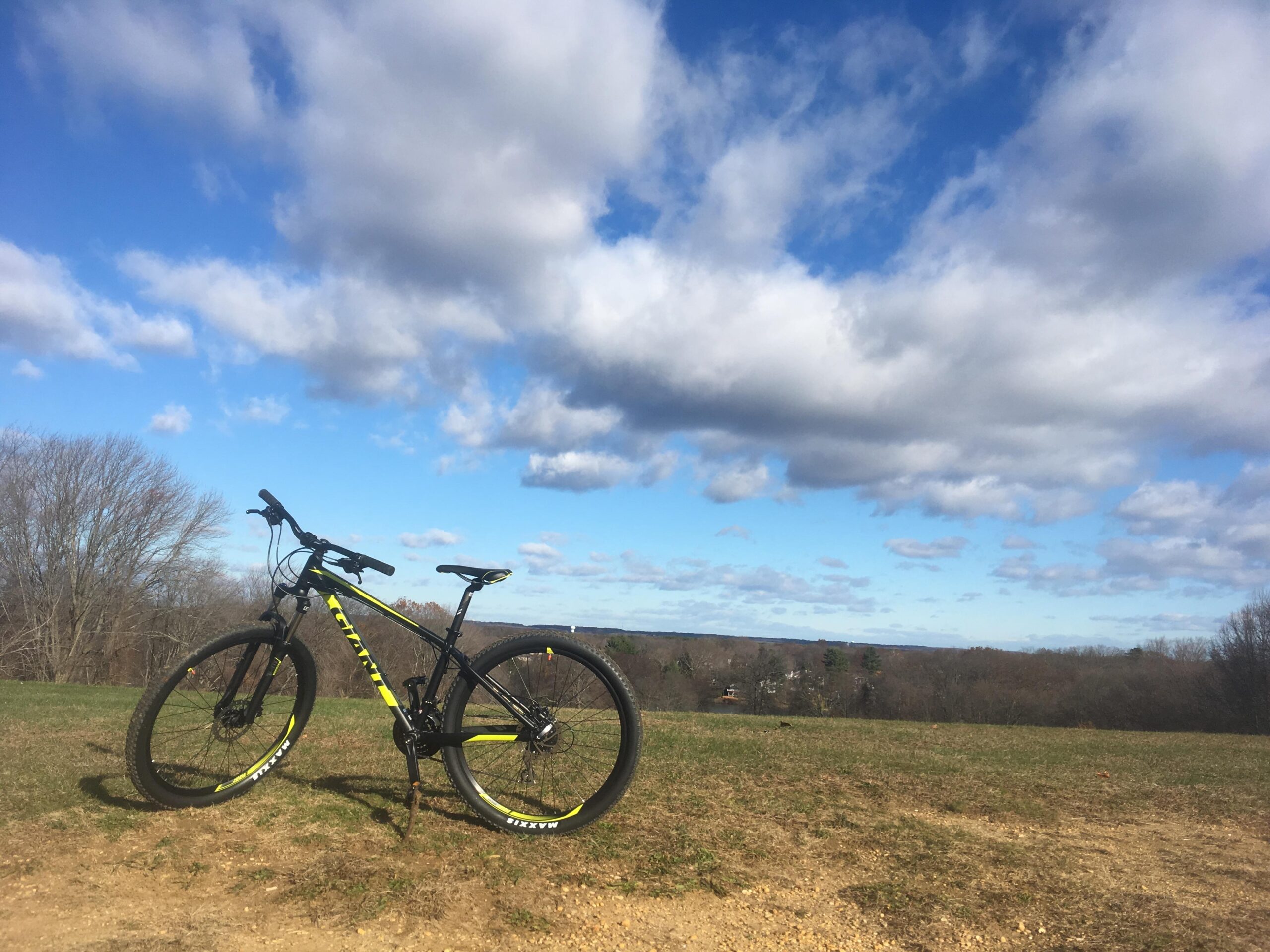 2017 Giant Talon 3: A black and yellow mountain bike resting on a grassy hill with a scenic view of rolling hills and a blue sky filled with fluffy clouds. The landscape appears serene, suggesting a perfect spot for outdoor biking or enjoying nature.