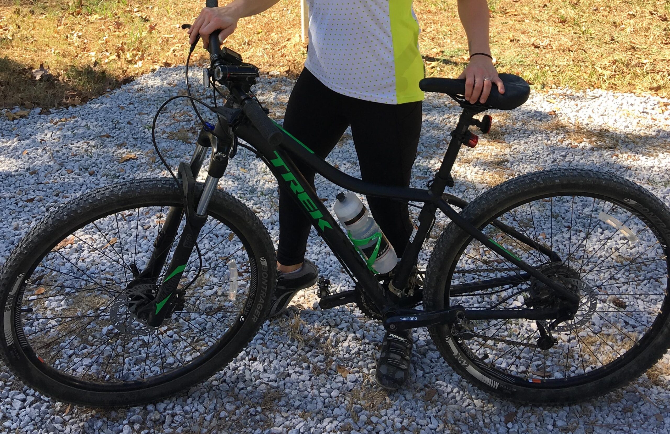 Trek Cali S: A woman stands next to a black mountain bicycle with green accents. She is wearing a bright yellow and white athletic shirt with polka dots, black leggings, and cycling shoes. A water bottle is attached to the bike frame. The background features a gravel path and some greenery.