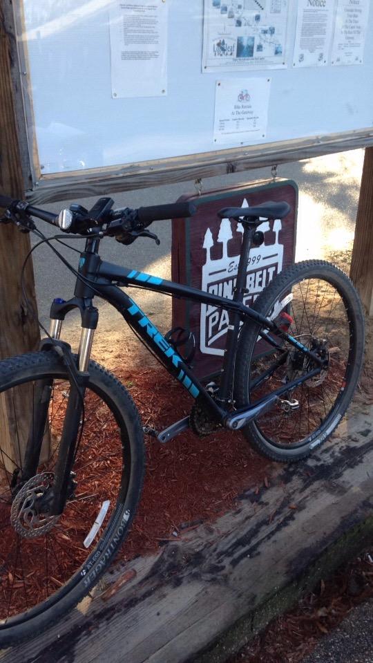 Trek X-Caliber 7: A mountain bike is parked beside a wooden sign displaying information and notices, set against a backdrop of red mulch. The bike features prominent suspension forks and a dark frame with blue accents.