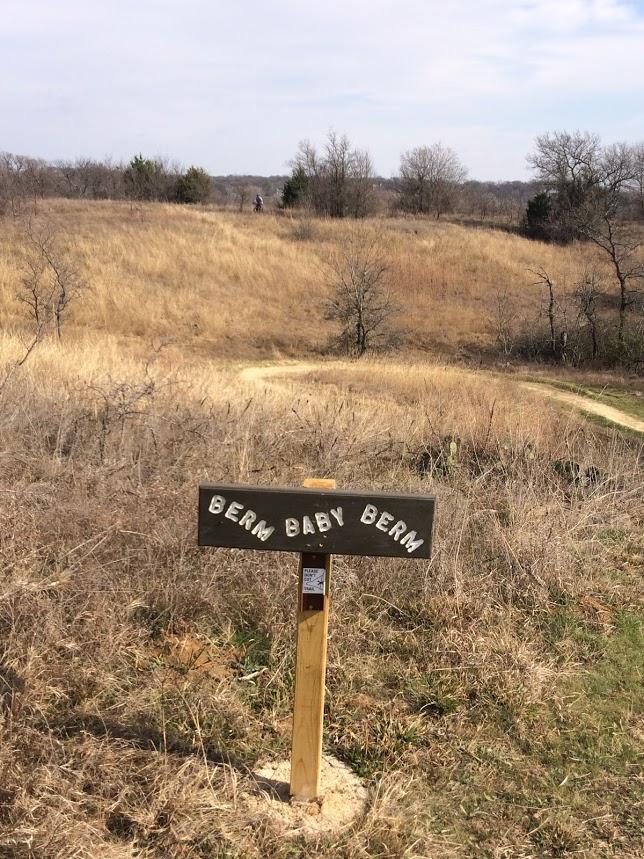A wooden sign reading "BERM BABY BERM" stands in the foreground of an open, grassy landscape on a sunny day, with a winding dirt path leading into the distance. Sparse trees dot the hillside, and a person is visible near the top of the hill, enhancing the natural setting. Knob Hills Grapevine Lake mountain bike trail.