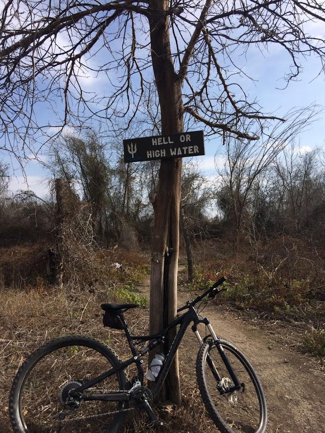 A mountain bike leaning against a tree with a sign that reads "HELL OR HIGH WATER." The background features sparse vegetation and a clear blue sky, indicating a natural outdoor setting. Knob Hills Grapevine Lake mountain bike trail.