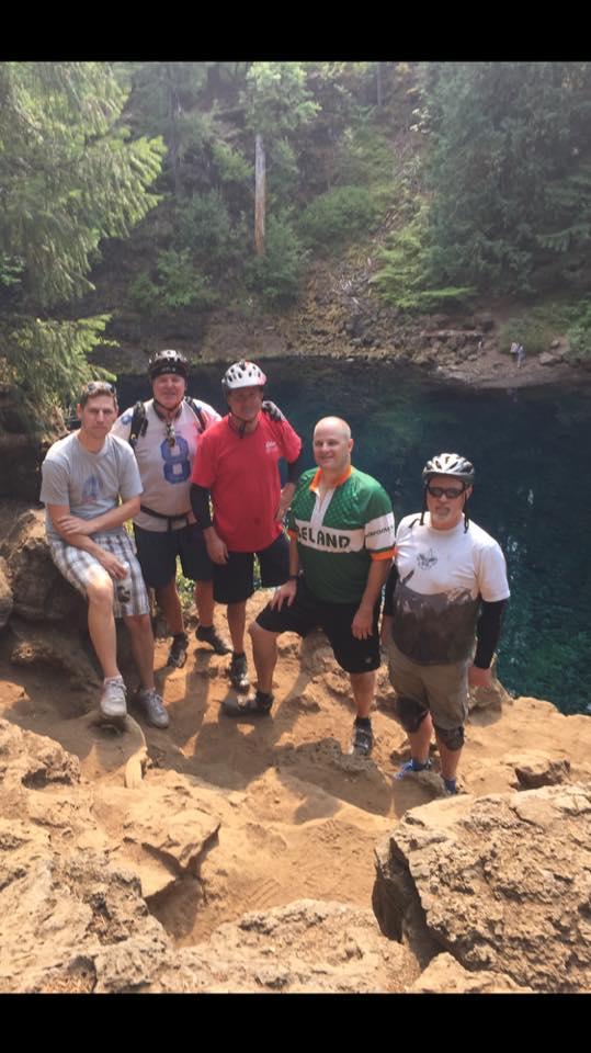 A group of five men posing on a rocky outcrop near a clear blue lake, surrounded by lush greenery. They are wearing casual clothing and helmets, indicating a cycling or outdoor adventure, with the landscape showcasing trees and a sandy area. Mckenzie River Trail mountain bike trail.
