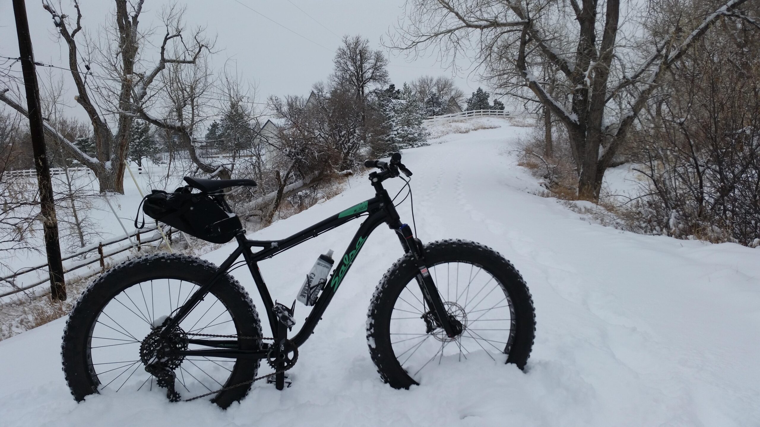 Salsa Mukluk: A fat tire bike parked in deep snow on a winter path, surrounded by leafless trees and a snow-covered landscape. The sky is overcast, adding to the wintry atmosphere.
