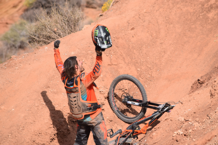 A mountain biker in an orange and black outfit celebrates with their helmet raised above their head, standing next to their bicycle that has fallen over on a dirt ramp. The background features a rugged, natural landscape.