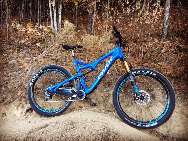 Pivot Trail 429: A blue mountain bike with a lightweight frame and wide tires, positioned on a dirt trail surrounded by autumn foliage. The bike features "Pivot" branding, a gold fork, and Maxxis tires.