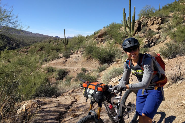 A mountain biker with a backpack smiles while resting beside her bike on a dirt trail surrounded by desert vegetation and cacti under a clear blue sky.