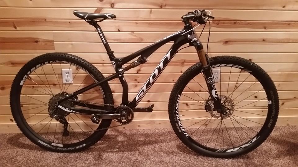 Scott Spark 900 Premium: A black mountain bike with the brand name "Scott" displayed prominently on the frame. It features a sleek design, front suspension, and rugged tires suitable for off-road trails. The bike is positioned upright against a wooden panel background, showcasing its detailed components and craftsmanship.