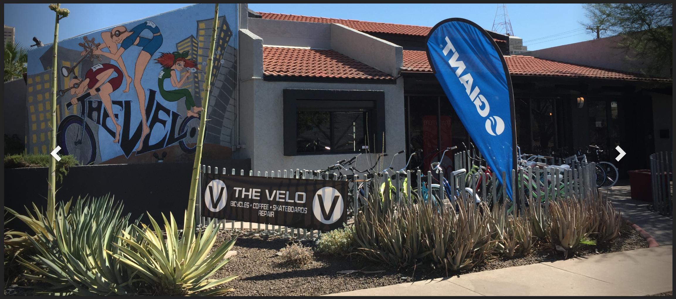 A vibrant mural depicting cyclists adorns the exterior of "The Velo," a bicycle shop featuring various bicycles displayed outside. The shop has a sign indicating its services, including bicycles, coffee, skateboards, and repairs. A large blue banner with the "Giant" logo flutters beside the entrance, while desert plants and landscaping frame the scene.
