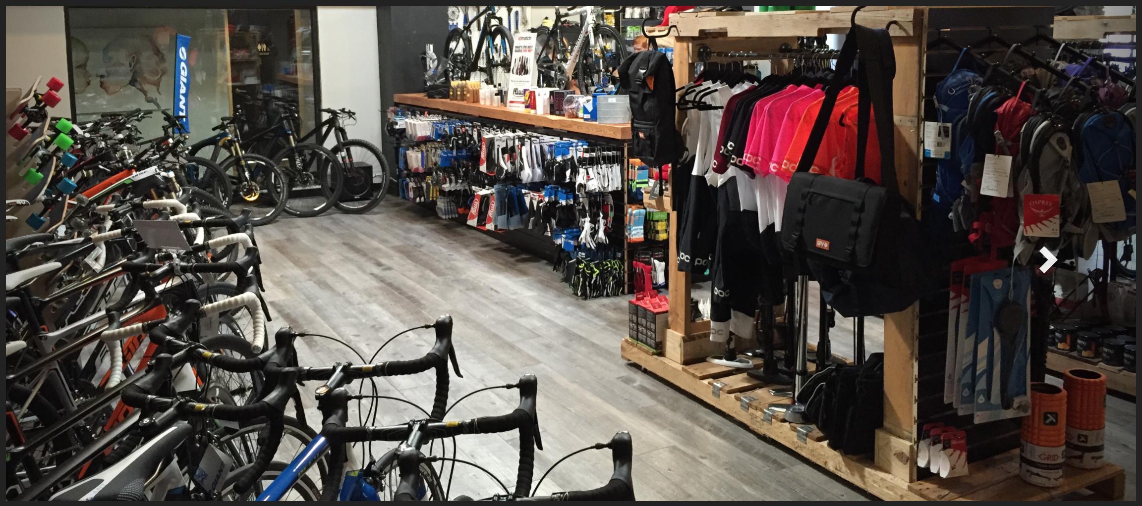 Alt text: Interior view of a bike shop showcasing a variety of bicycles and cycling gear. The shop features rows of bikes, including road and mountain bikes, alongside shelves stocked with clothing, accessories, and equipment for cyclists. The flooring is wooden, contributing to a modern retail atmosphere.