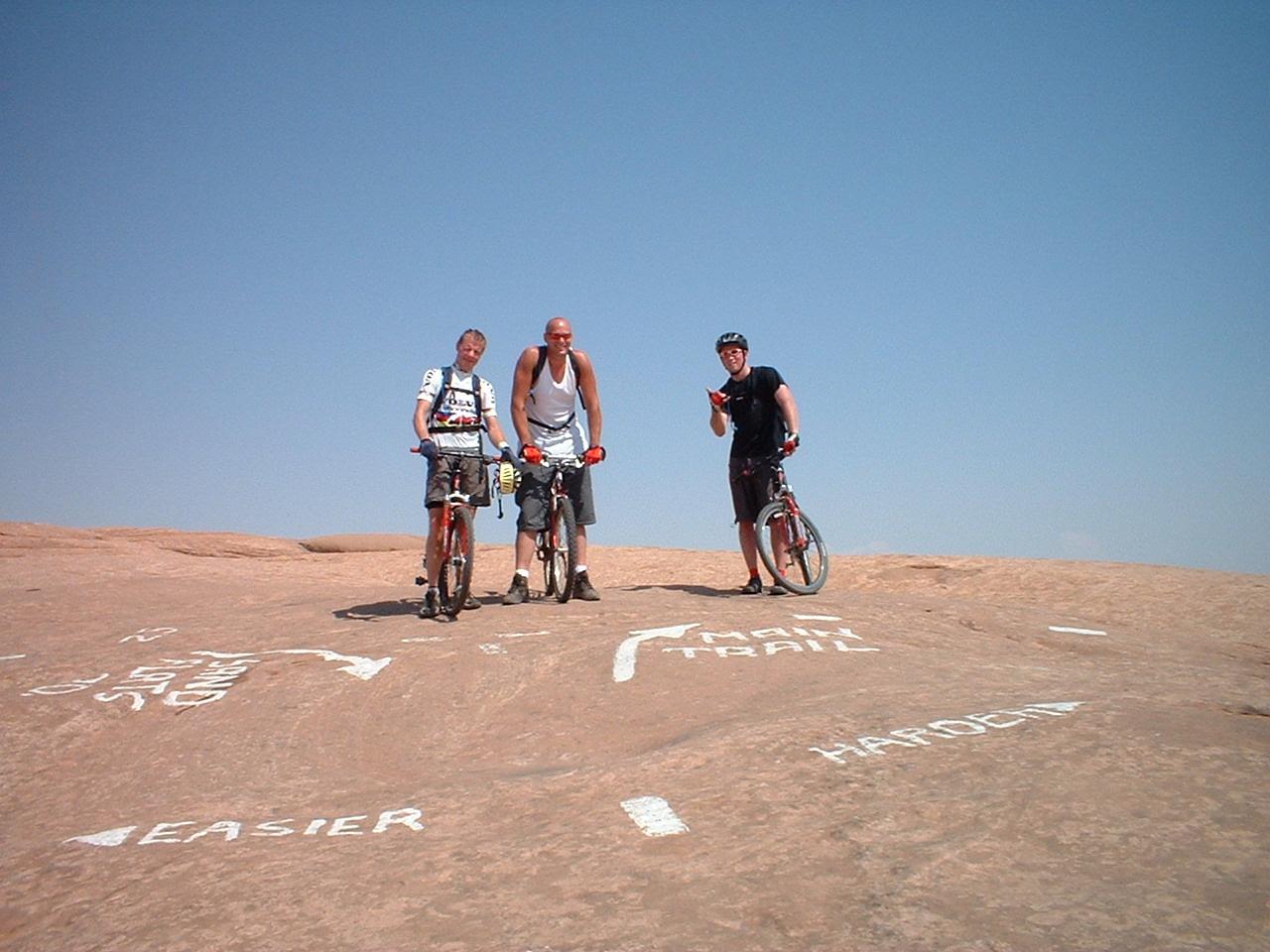Three mountain bikers pose on a rocky terrain under a clear blue sky. Two riders stand beside their bicycles, while the third rider gives a thumbs-up gesture. The ground is marked with directional arrows and text indicating trail difficulty levels labeled "EASIER," "MAIN TRAIL," and "HARDCORE." Slickrock mountain bike trail.