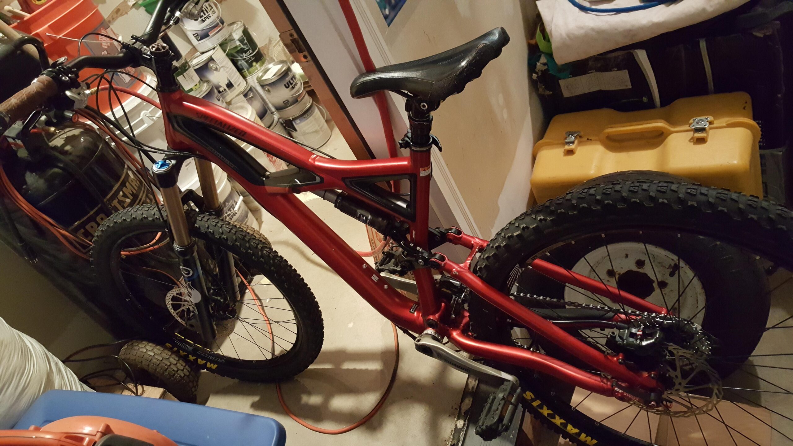 Specialized Enduro Expert: A red mountain bike is propped against a wall in a garage, with its front wheel partially turned. Various items, including cans of paint and a yellow toolbox, are visible in the background. The bike has thick tires and is equipped with a suspension fork and a dropper seat post.