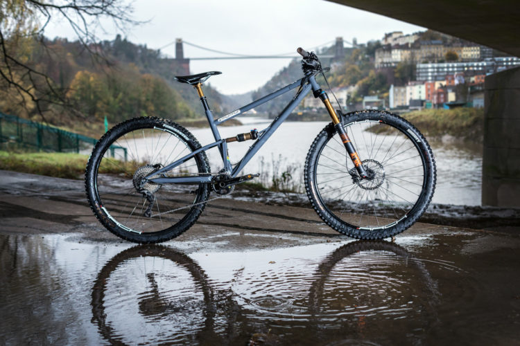 A mountain bike standing on a wet surface with a reflection in a puddle. In the background, there is a river and a bridge, surrounded by green trees and buildings, creating a scenic outdoor setting. The weather appears overcast, suggesting a damp atmosphere.